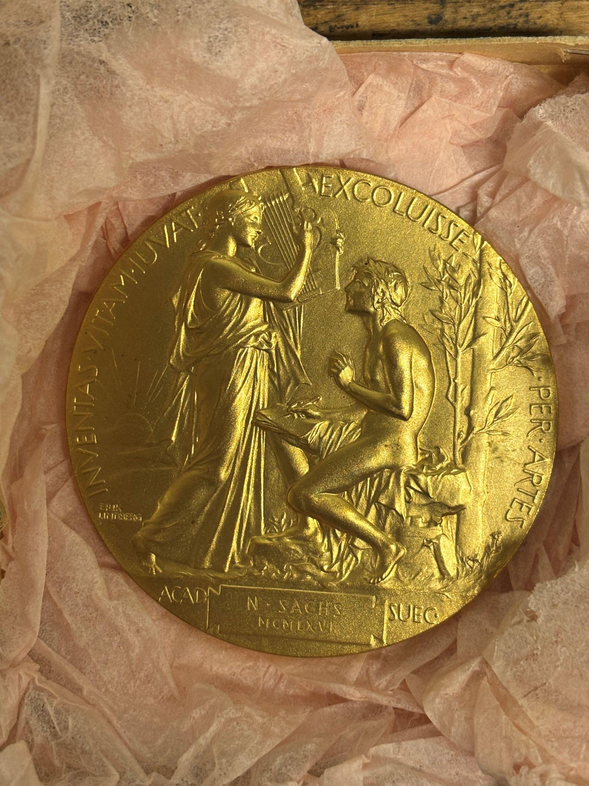 Guldfärgad medalj föreställande en stående kvinna och en sittande man i relief. Kvinnan håller upp en lyra, mannen håller i skrivdon. Runt medaljen löper en text i latin och i nedre delen syns namnet N. Sachs.
